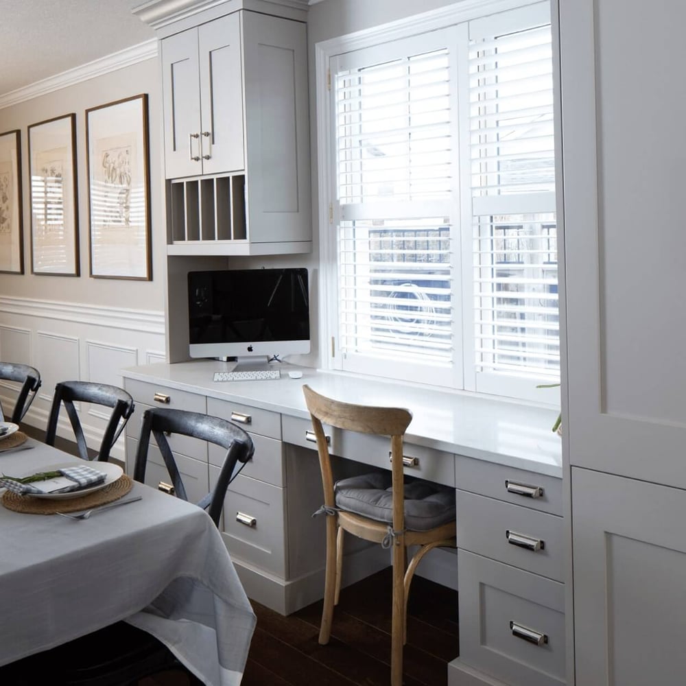 Project_Kitchens_Eastbridge12 Modern kitchen interior featuring a long dining table, gray cabinetry, and a built-in workspace with a computer and window shutters.