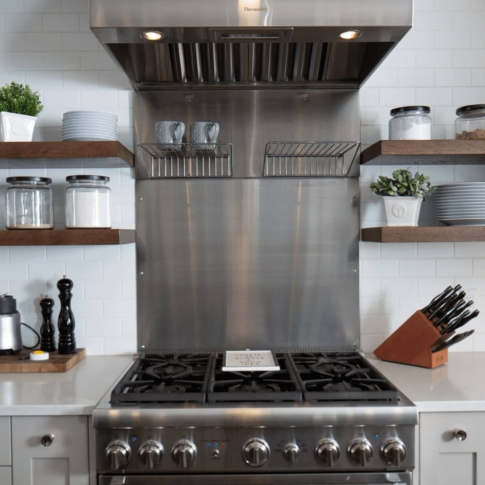 Project_Kitchens_Eastbridge6 Stainless steel kitchen range with a hood, open shelves displaying glass jars and plates, and a knife block on a countertop.