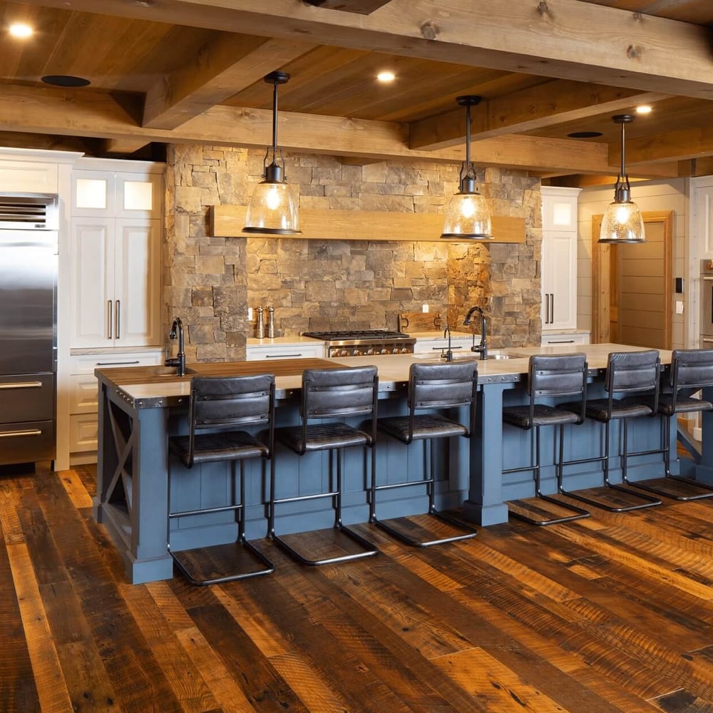 Project_Kitchens_Fruit+Ridge2 Modern kitchen with a large island, wooden beams, stone accents, and stainless steel appliances, featuring bar stools along the island.