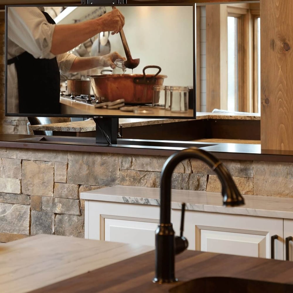 Project_Kitchens_Fruit+Ridge6 Kitchen interior with a television displaying a person stirring a pot, featuring wooden cabinetry and a stone countertop.