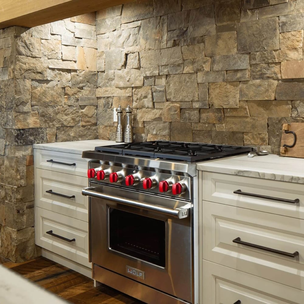 Project_Kitchens_Fruit+Ridge8 Stainless steel gas range with red knobs installed in a kitchen with stone wall and white cabinetry.