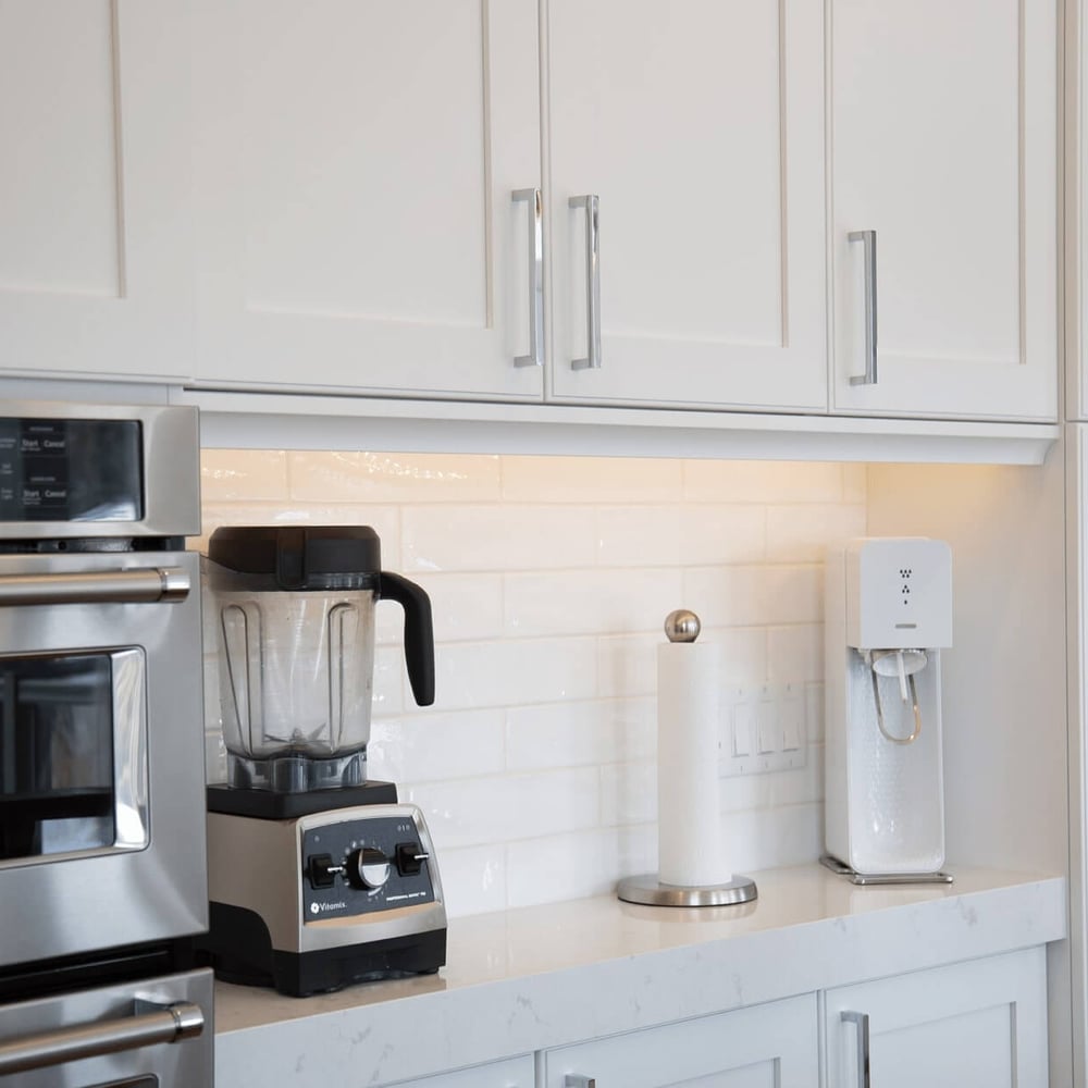 Modern kitchen featuring stainless steel appliances, a blender, and a water dispenser against white cabinetry and a tiled backsplash.