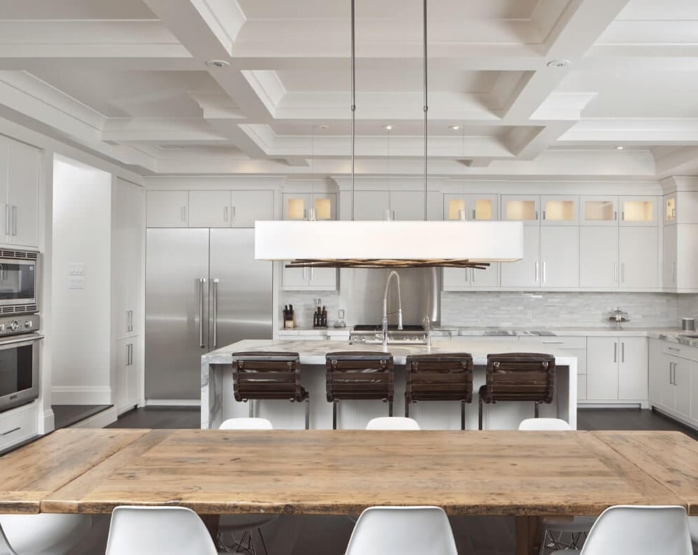 Project_Kitchens_Old+Colonial+Acres_MAIN Modern kitchen with white cabinetry, stainless steel appliances, and a wooden dining table with white chairs.