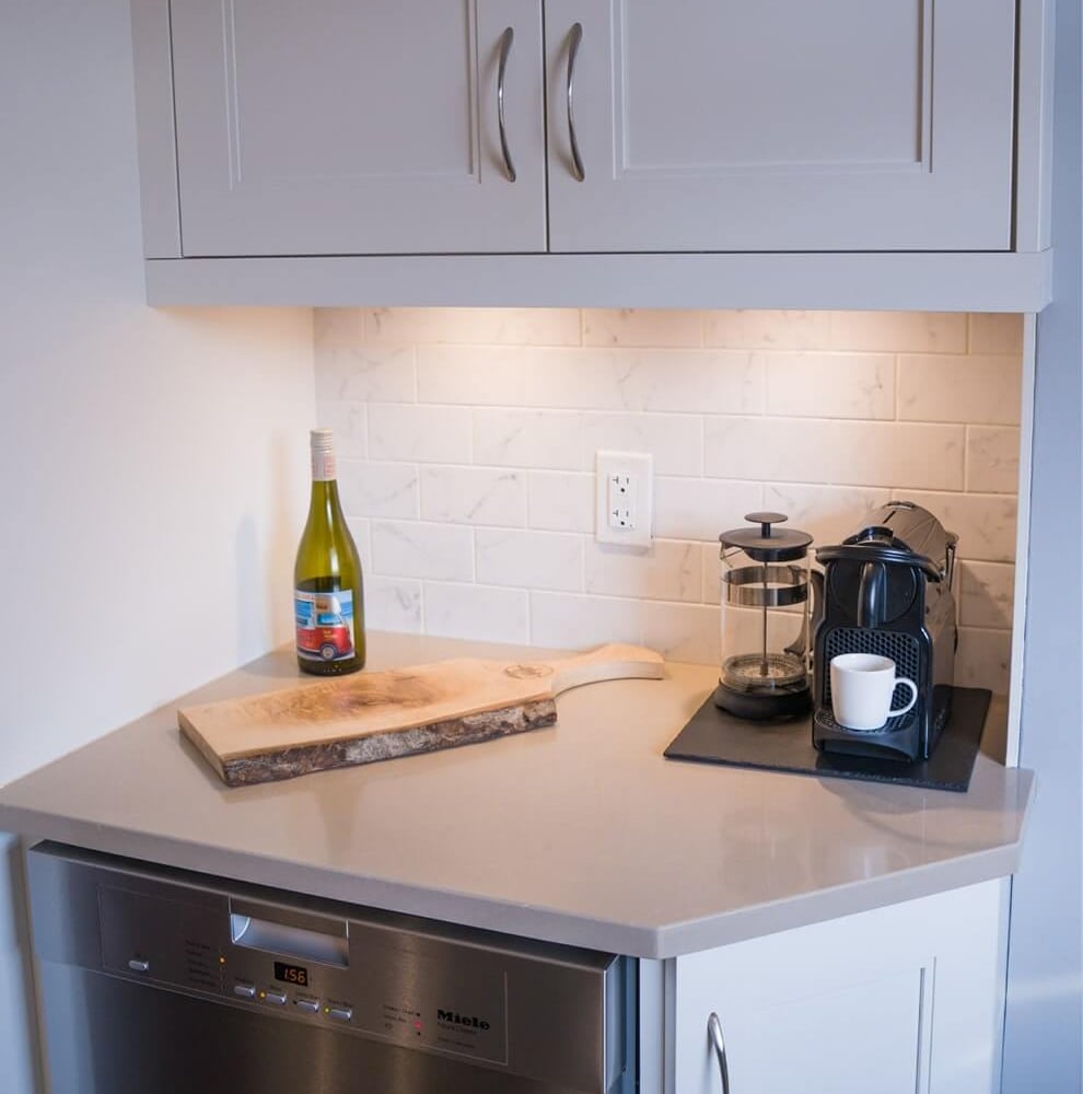 Project_Kitchens_Old+South12 Old South Kitchen area with white shaker cabinets, walnut floating shelves, under-cabinet lighting, a small wine bottle, coffee station, and a Miele dishwasher beneath a light countertop.