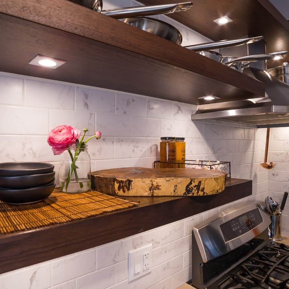 Project_Kitchens_Old+South5 OldSouth kitchen scene with white marble tile backsplash, dark wood open shelves, a stainless range hood, a round wooden cutting board, and a glass vase with pink ranunculus alongside spice jars.