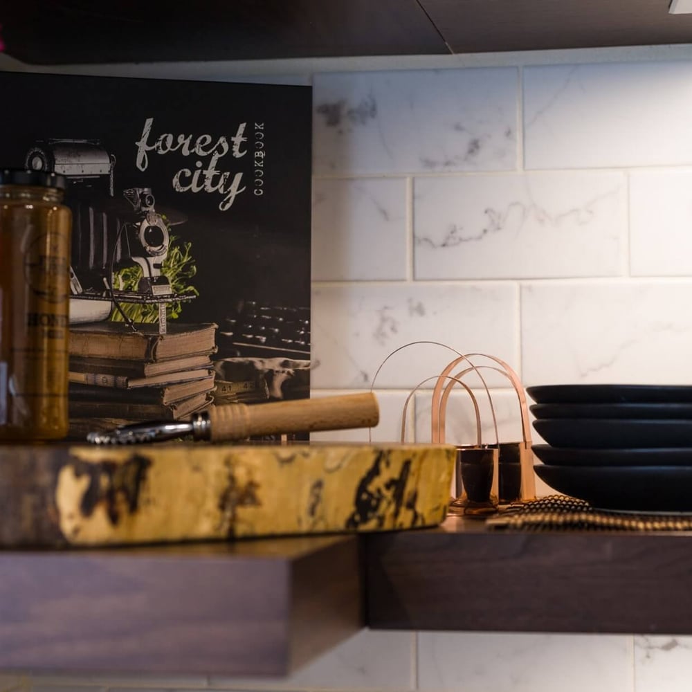Project_Kitchens_Old+South7 Old South Kitchen display with a dark walnut shelf, copper utensil accents, stacked black plates, glass bottle and a chalkboard-style forest city cookbook against a white marble tile backsplash