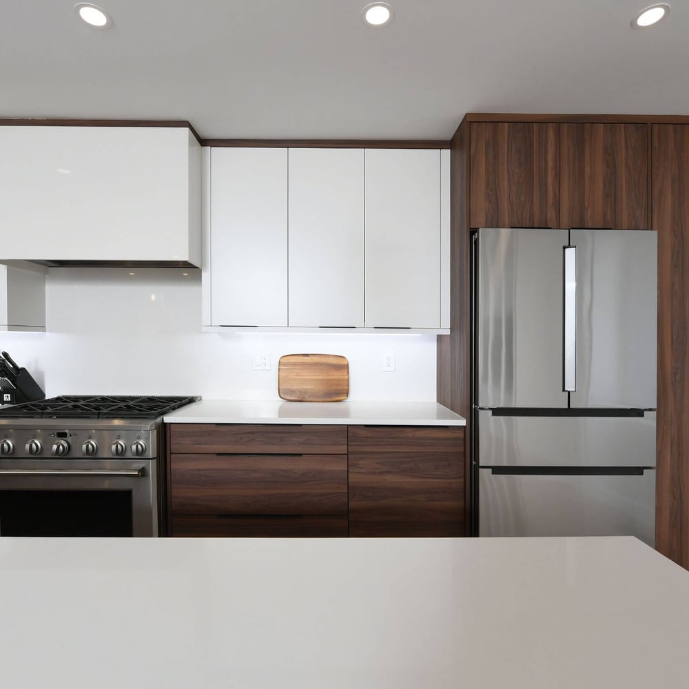 Modern kitchen with white upper cabinets and dark wood lower cabinets, stainless steel refrigerator and range, white countertops, and a wooden cutting board on the counter
