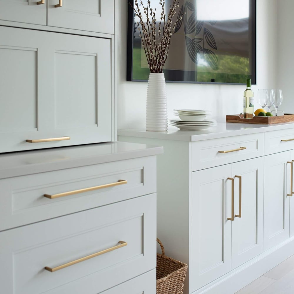 White shaker kitchen cabinetry with brass handles, a walnut-toned cabinet, and a bright window beside styled decor.