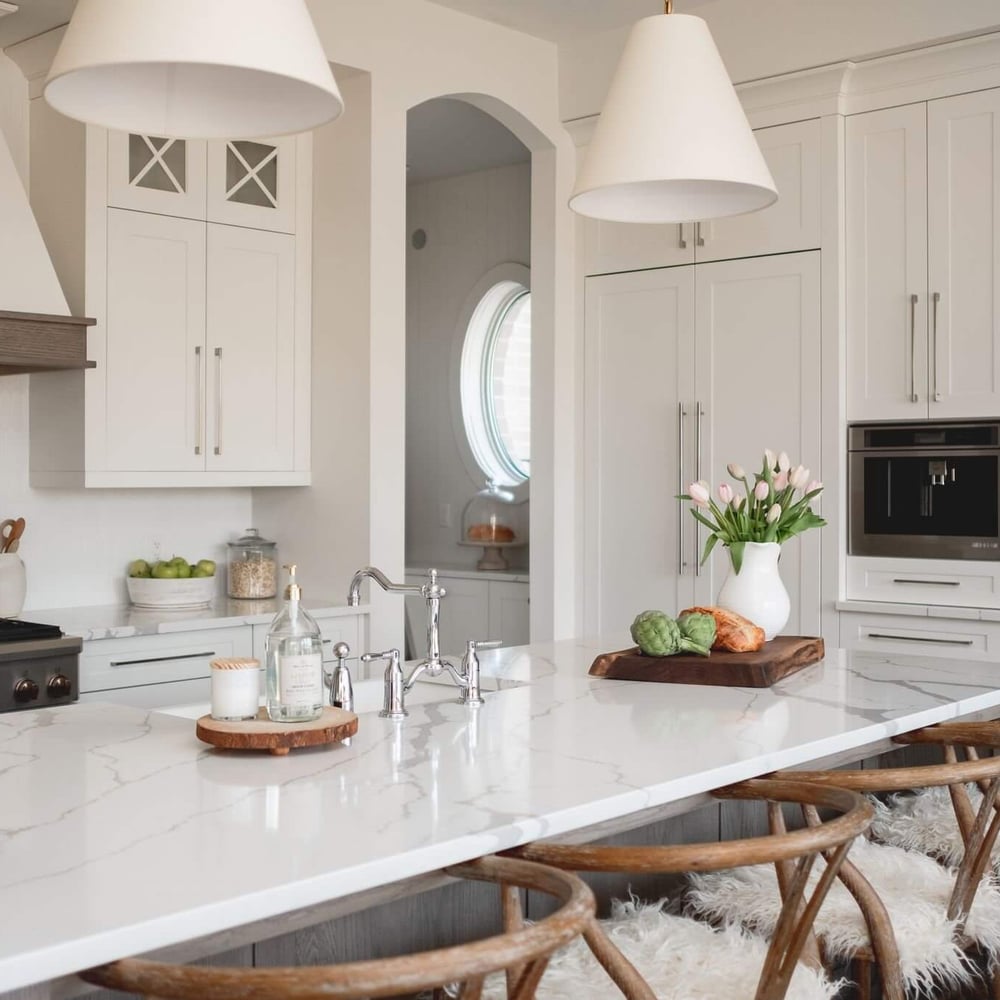 White modern farmhouse kitchen with a marble island, white cabinetry, and wooden bar stools, featured in Byron Beauty by Casey's Kitchens in London.