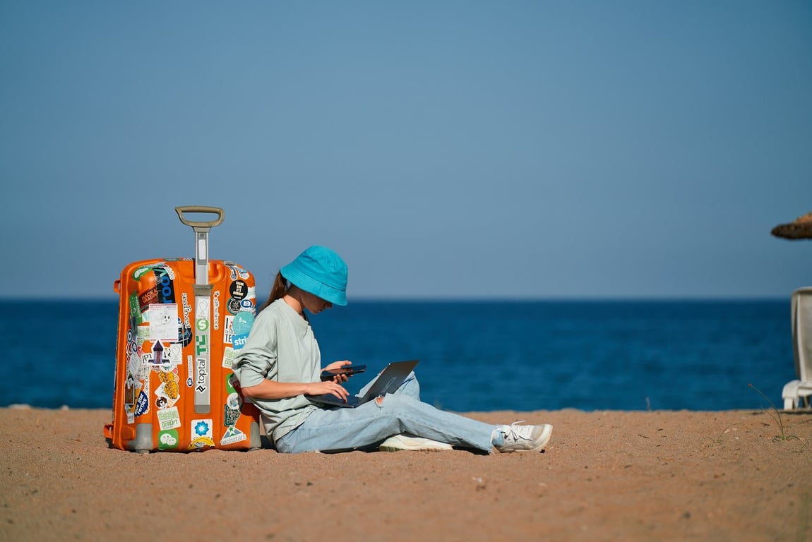 A girl sitting on the beach working on her laptop with her smartphone and luggage
