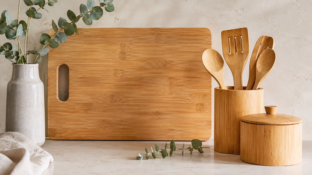 A wooden cutting board with bamboo kitchen utensils and a vase with greenery on a countertop.