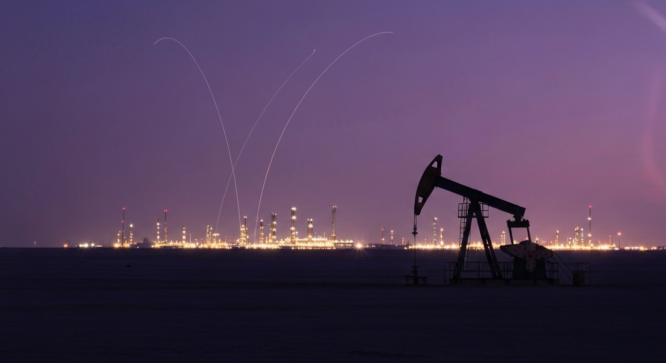 Oil pumpjack silhouette on a desert horizon at dusk with distant refinery lights and subtle upward light trails