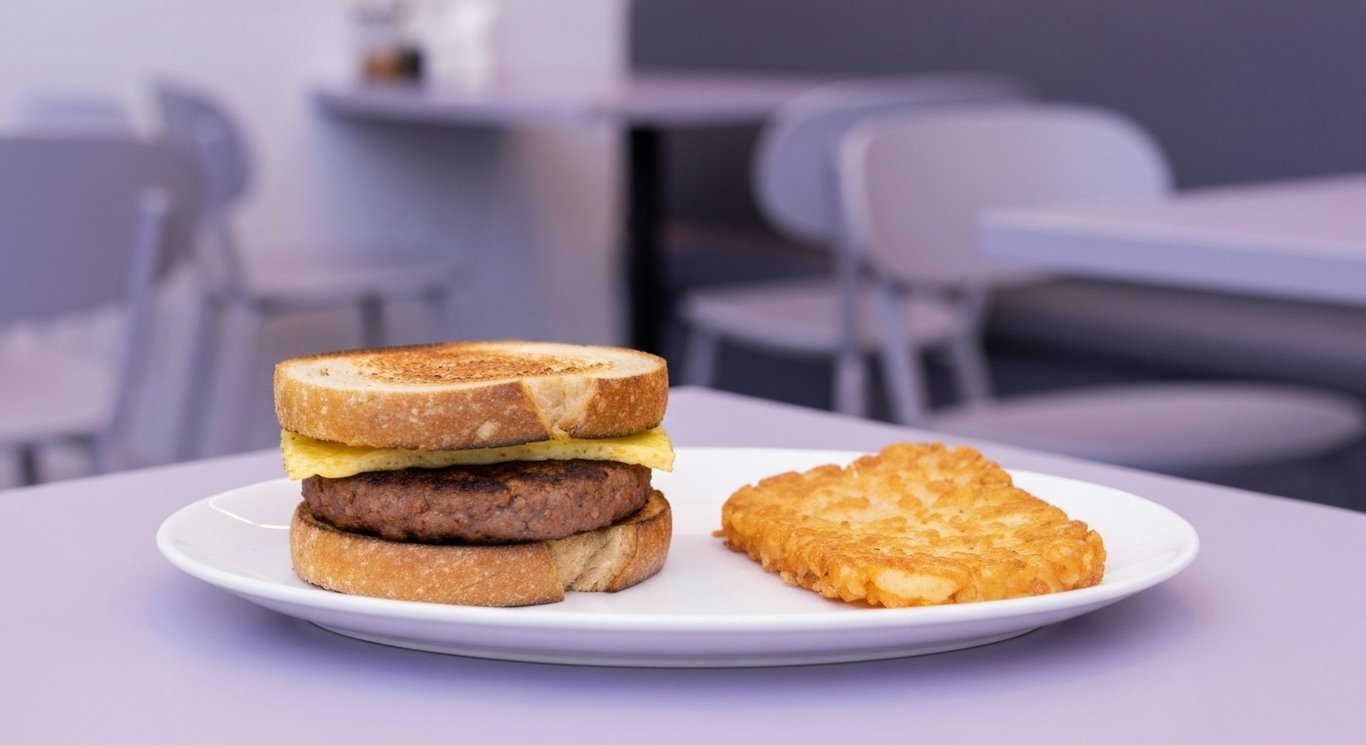 Breakfast sandwich with spicy patty on a plate beside hashbrowns in a modern café with soft purple hues
