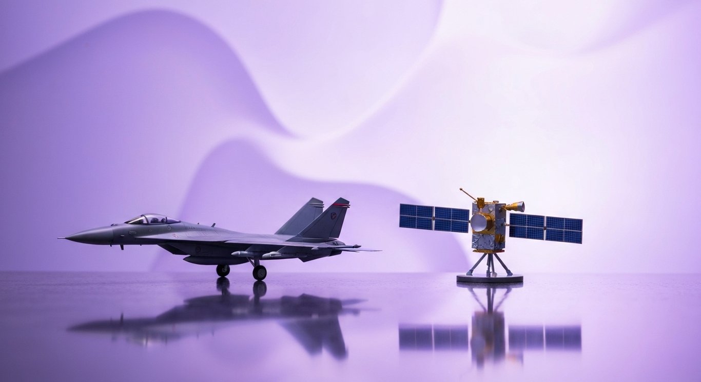Modern fighter jet and satellite models displayed on a reflective surface with a soft purple background