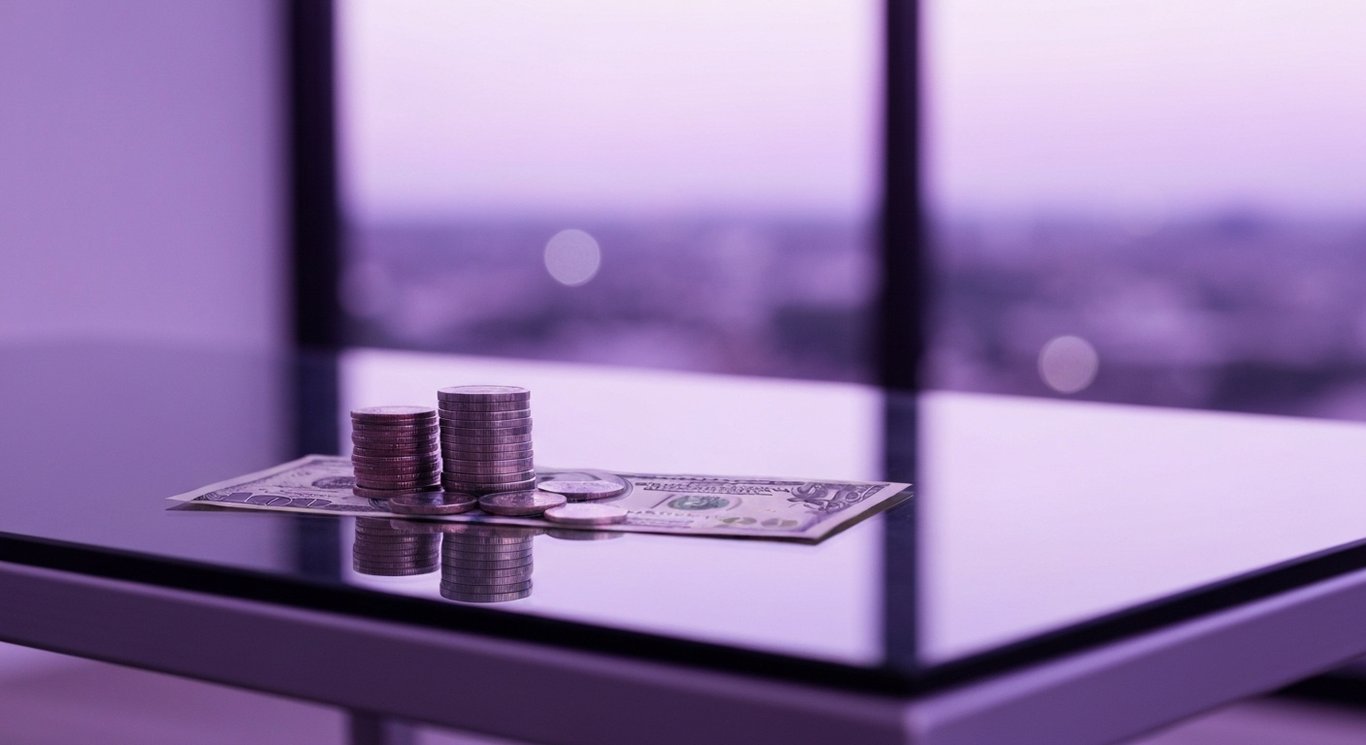 Stack of coins and dollar bills on a glass table with a blurred purple cityscape in the background