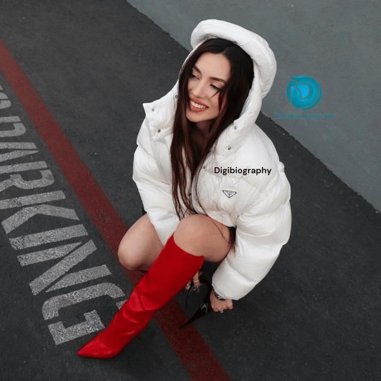 Ava Max sitting on the road and wearing a white jacket with red boots