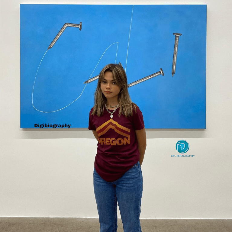 Ariana Greenblatt stands in front of the blue board while wearing a marron color
