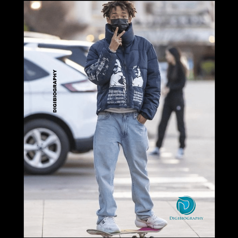 Jaden Smith stands on the skateboard while wearing a blue jacket and light blue jeans 