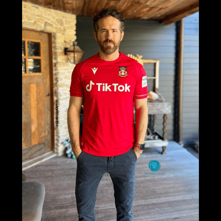 Ryan Reynolds stands in her house while wearing a red t-shirt and grey jeans