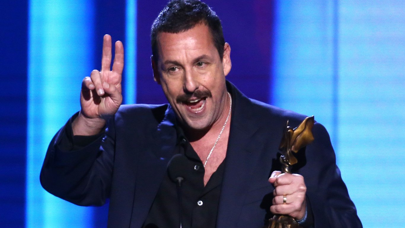 Adam Sandler stands on the stage holding an award in his hand and wearing a blue and black suit