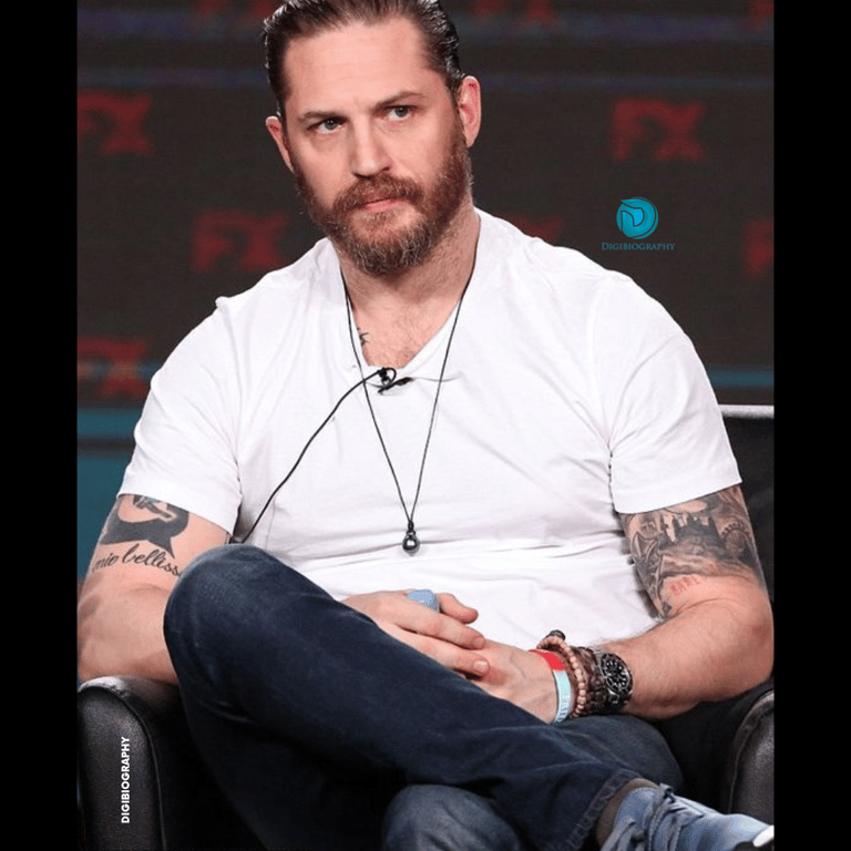Tom Hardy sitting on the sofa while wearing a white t-shirt and blue jeans