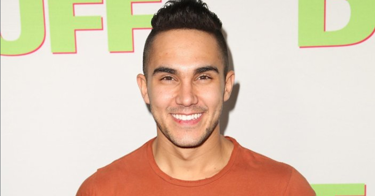 Carlos PenaVega wears a orange t-shirt and stand in front of award banner 