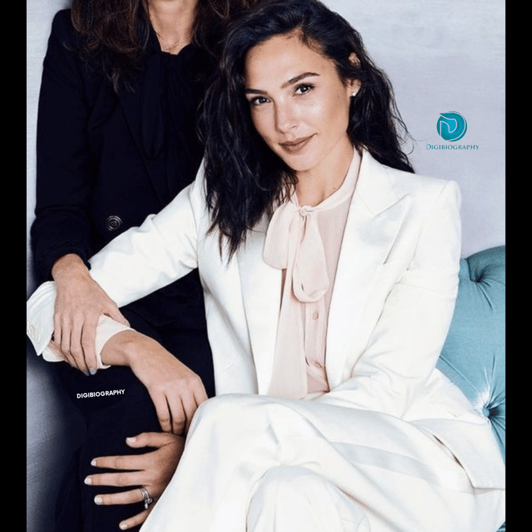 Gal Gadot wearing a white blazer and pink dress while sitting on the chair