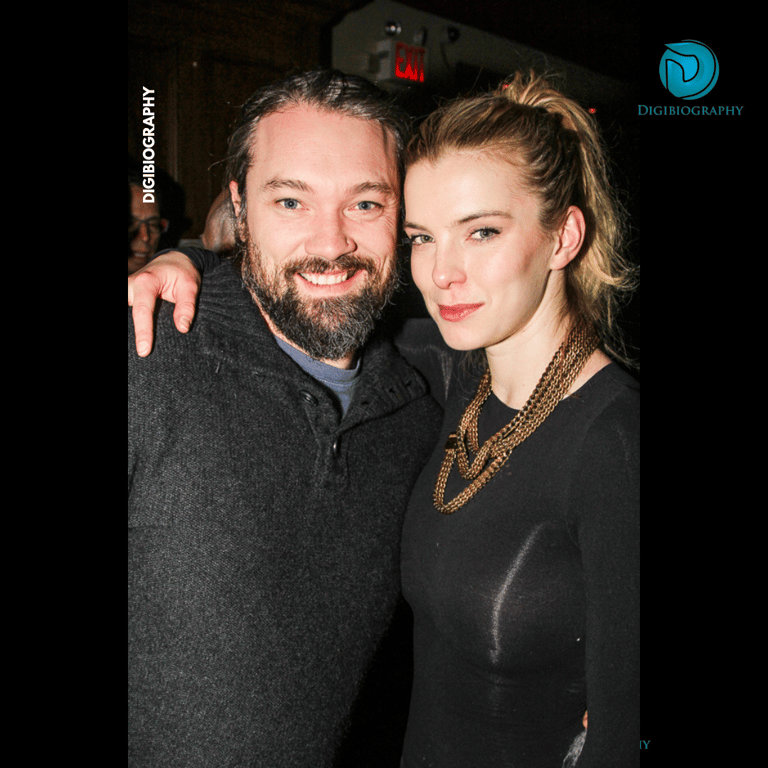 Betty Gilpin stands with her husband Cosmo Pfeil and wearing a black dress