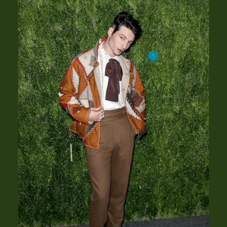 Ezra Miller stands in the green background and wearing a orange with a white jacket