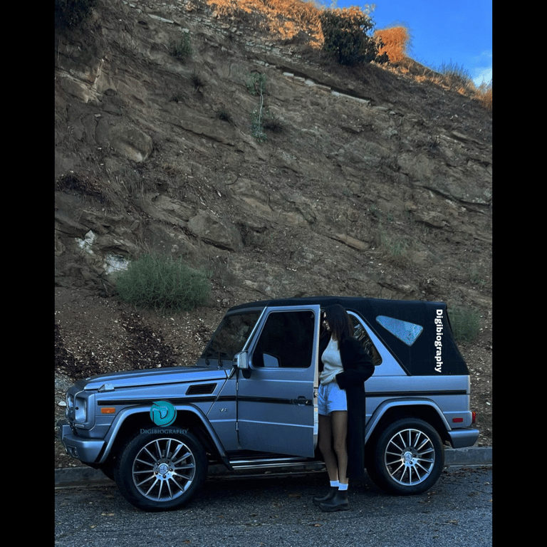 Kendall Jenner wearing a white top and black long coat stands beside the car on the road