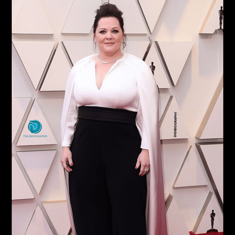 Melissa McCarthy stands close to the wall while wearing a white and black dress