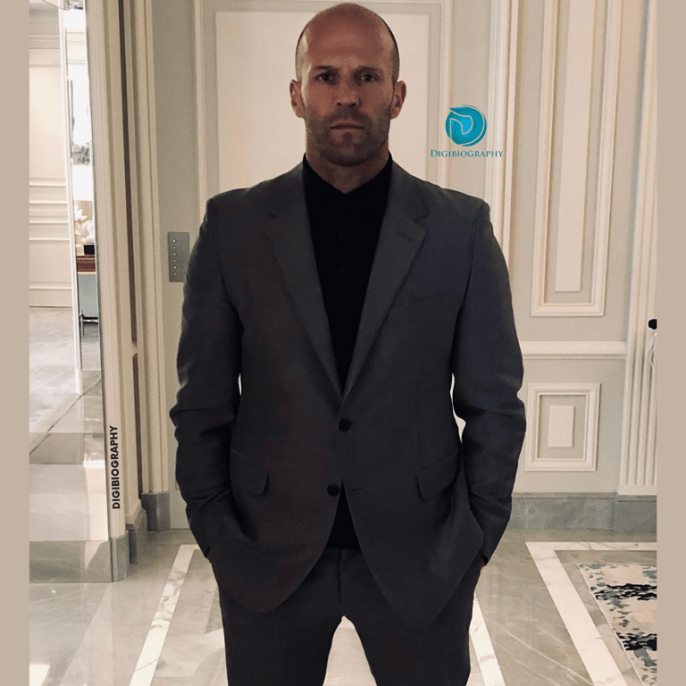 Jason Statham stands in her house while wearing a gray coat