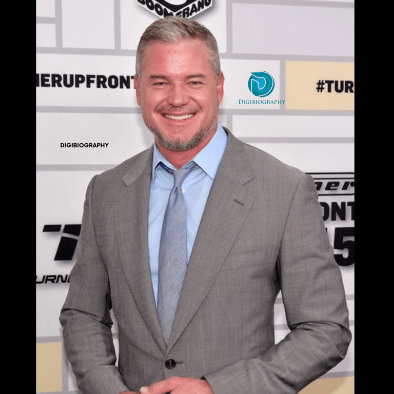Eric Dane wearing a grey coat with a blue shirt and gives a smile