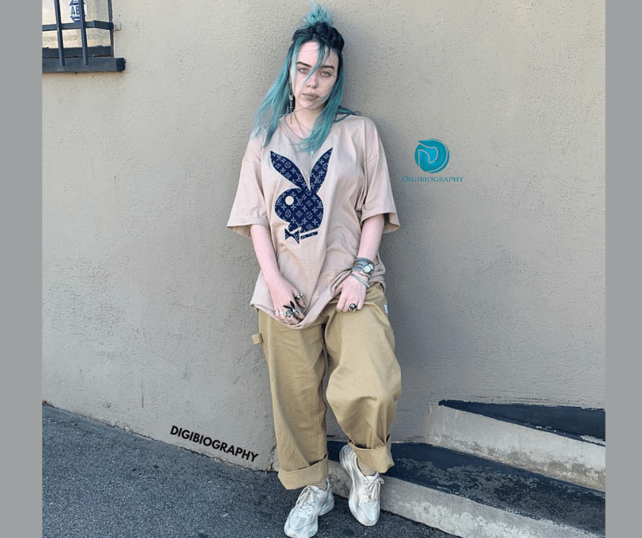 Billie Eilish wearing a light pink color t-shirt and stand close to the wall