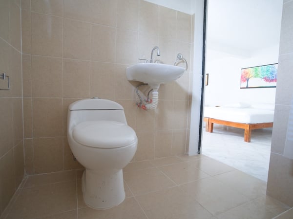 Baño privado con azulejo beige, lavabo y sanitario blancos; al fondo se aprecia la recámara con cama de madera