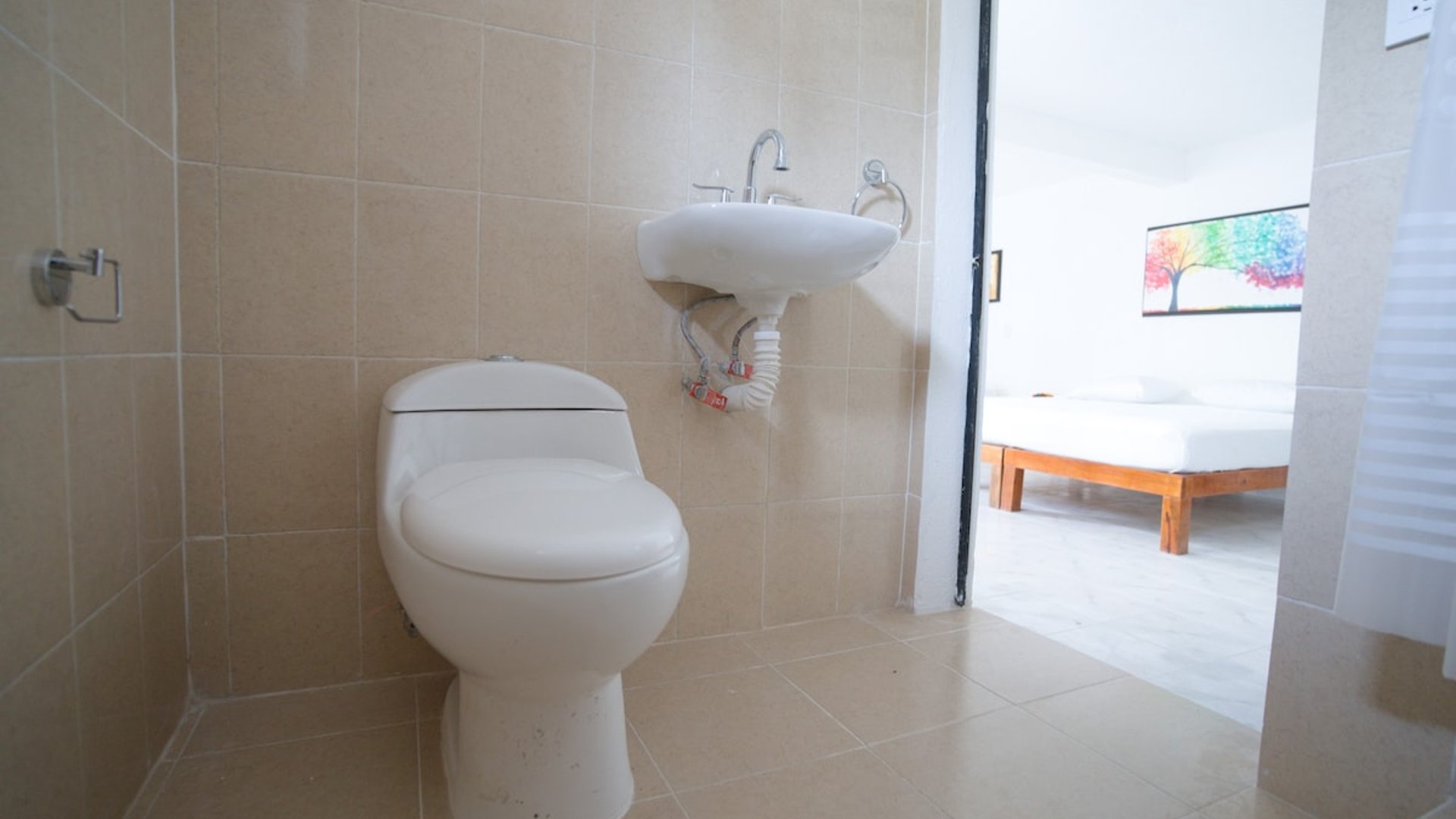Baño privado con azulejo beige, lavabo y sanitario blancos; al fondo se aprecia la recámara con cama de madera