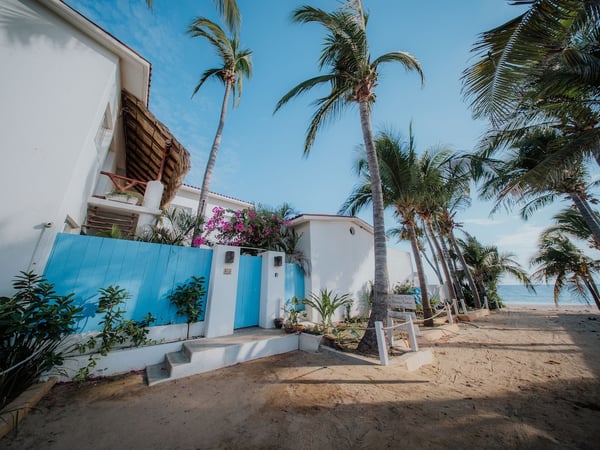 Fachada blanca con portón azul y bugambilias rosas frente a la playa, rodeada de palmeras y mar al fondo