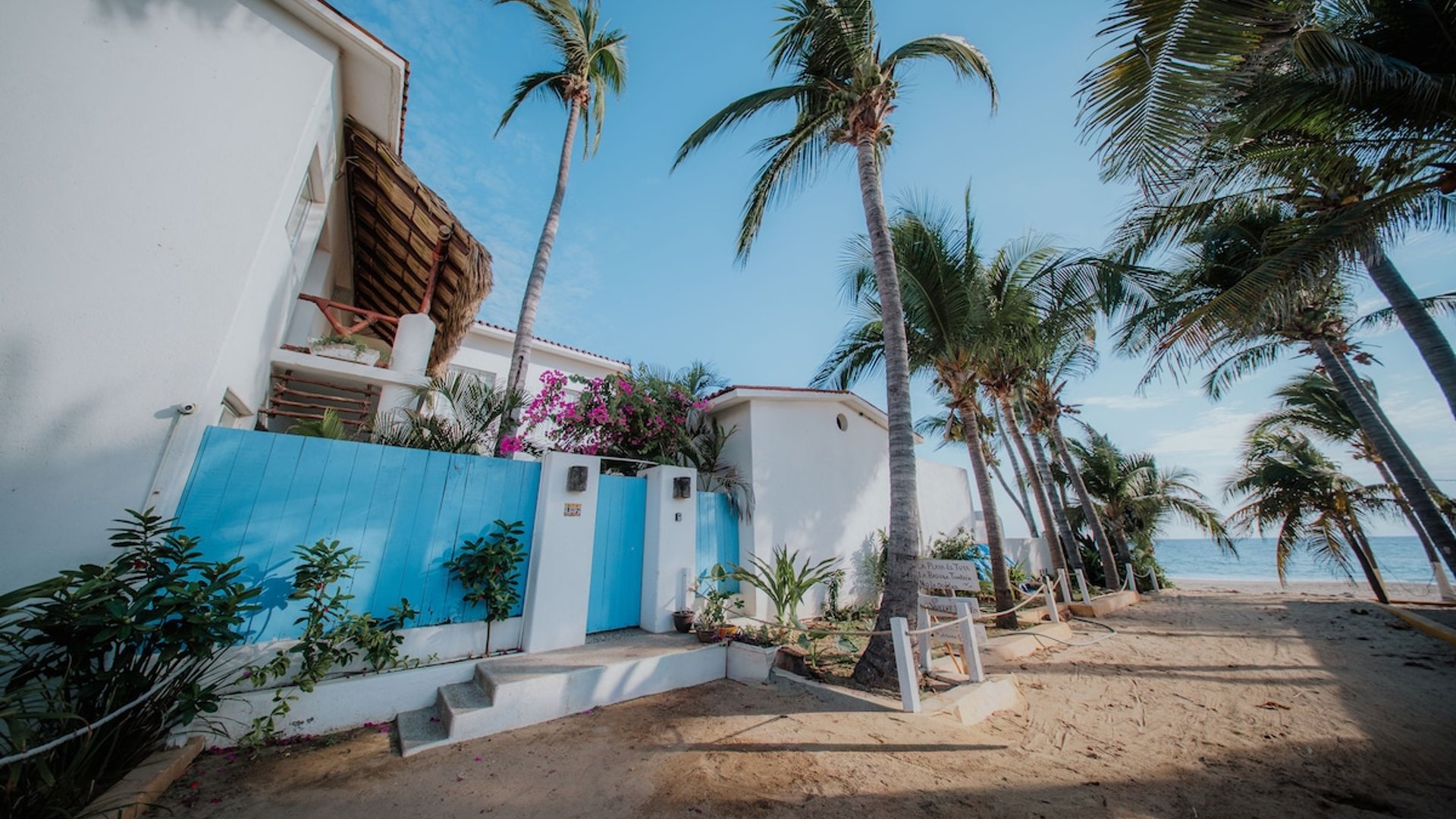 Fachada blanca con portón azul y bugambilias rosas frente a la playa, rodeada de palmeras y mar al fondo