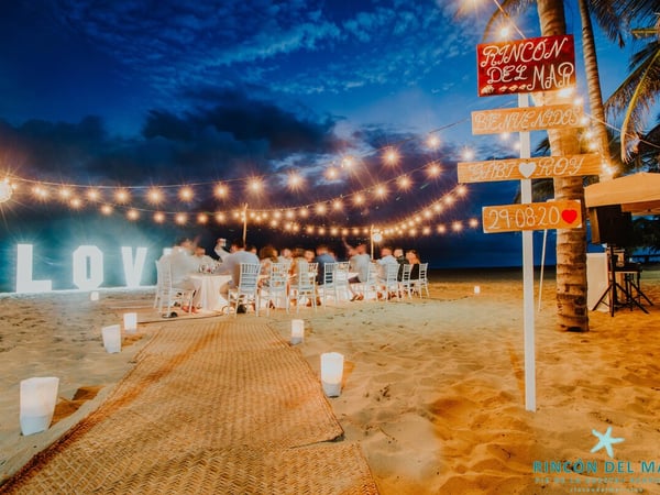 Boda en la playa al anochecer con luces colgantes, letras LOVE iluminadas y mesas de banquete frente al mar