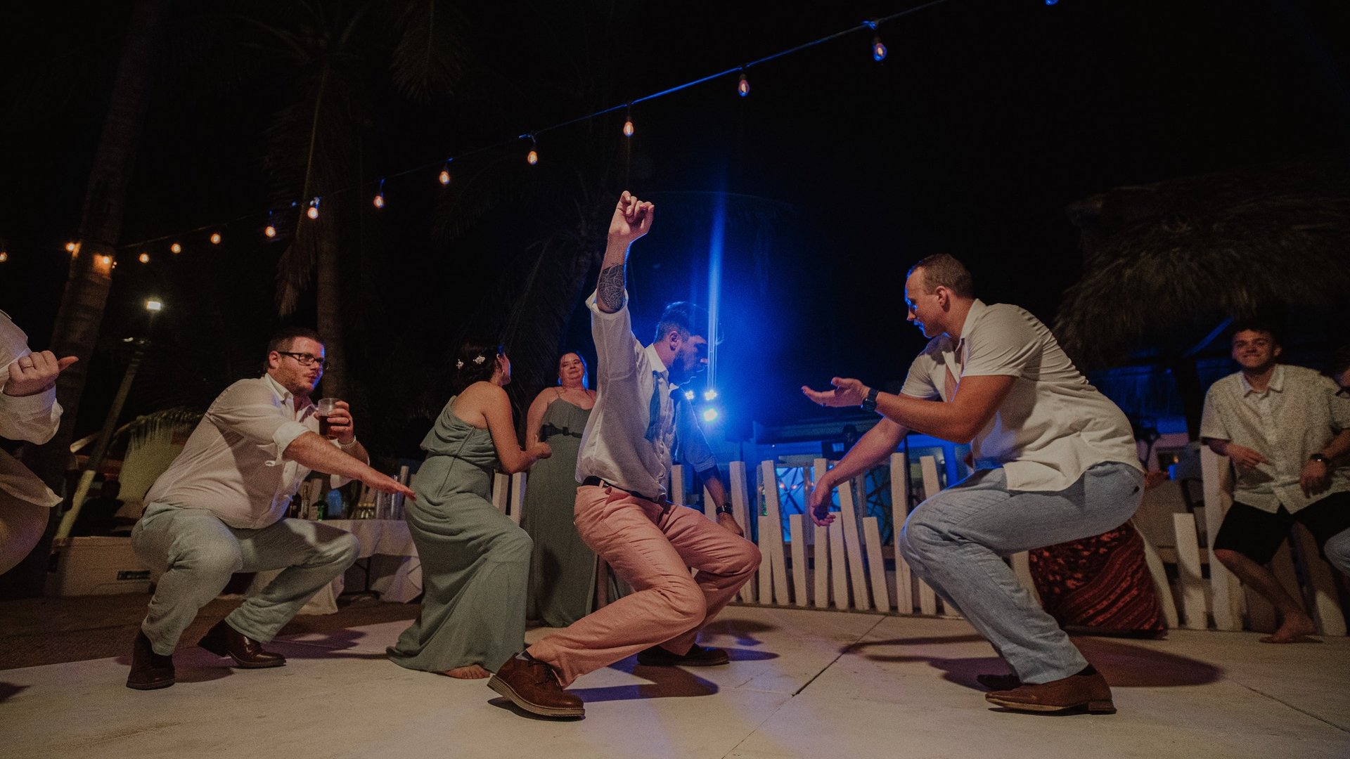 Invitados bailando en pista nocturna con luces colgantes y palmeras de fondo