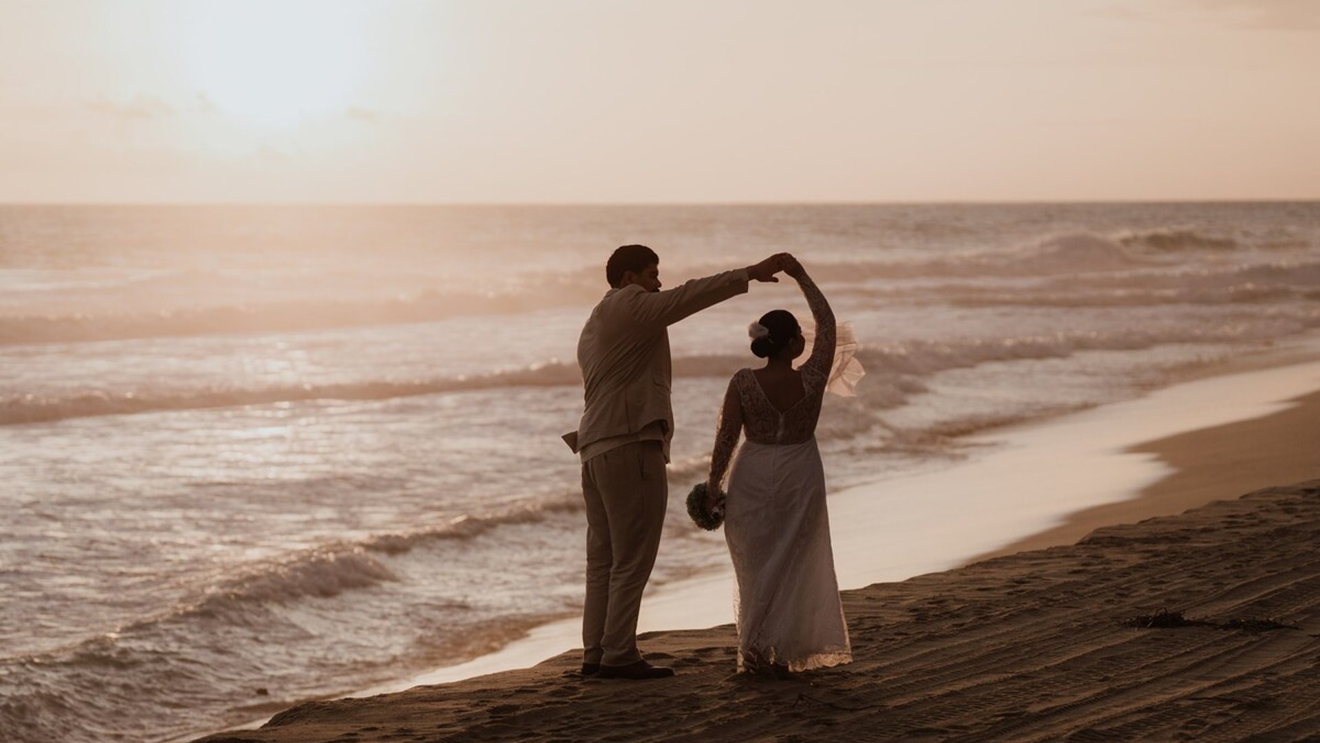Pareja de novios bailando en la playa al atardecer frente al mar Pacífico con luz dorada