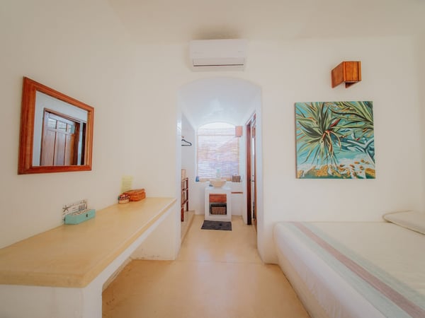 Recámara blanca con tocador empotrado, espejo de madera, aire acondicionado y cuadro tropical; pasillo arqueado al fondo
