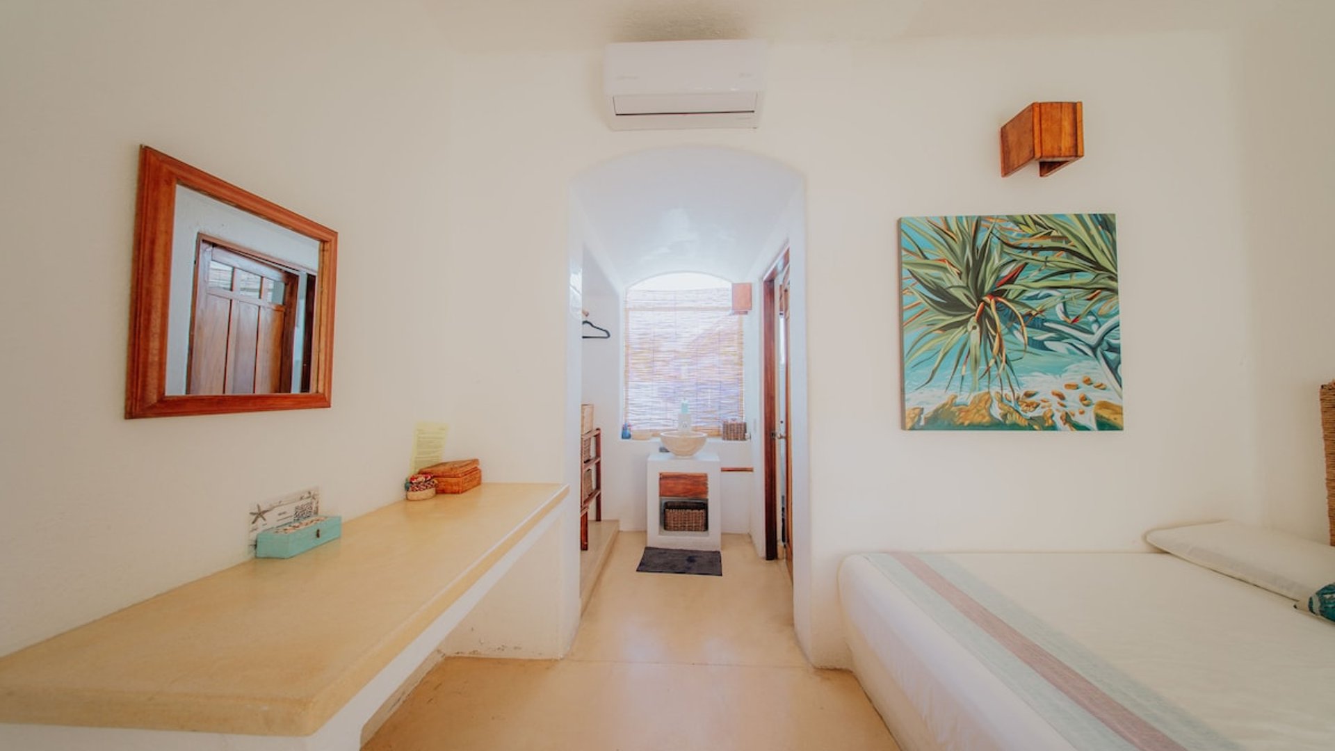 Recámara blanca con tocador empotrado, espejo de madera, aire acondicionado y cuadro tropical; pasillo arqueado al fondo