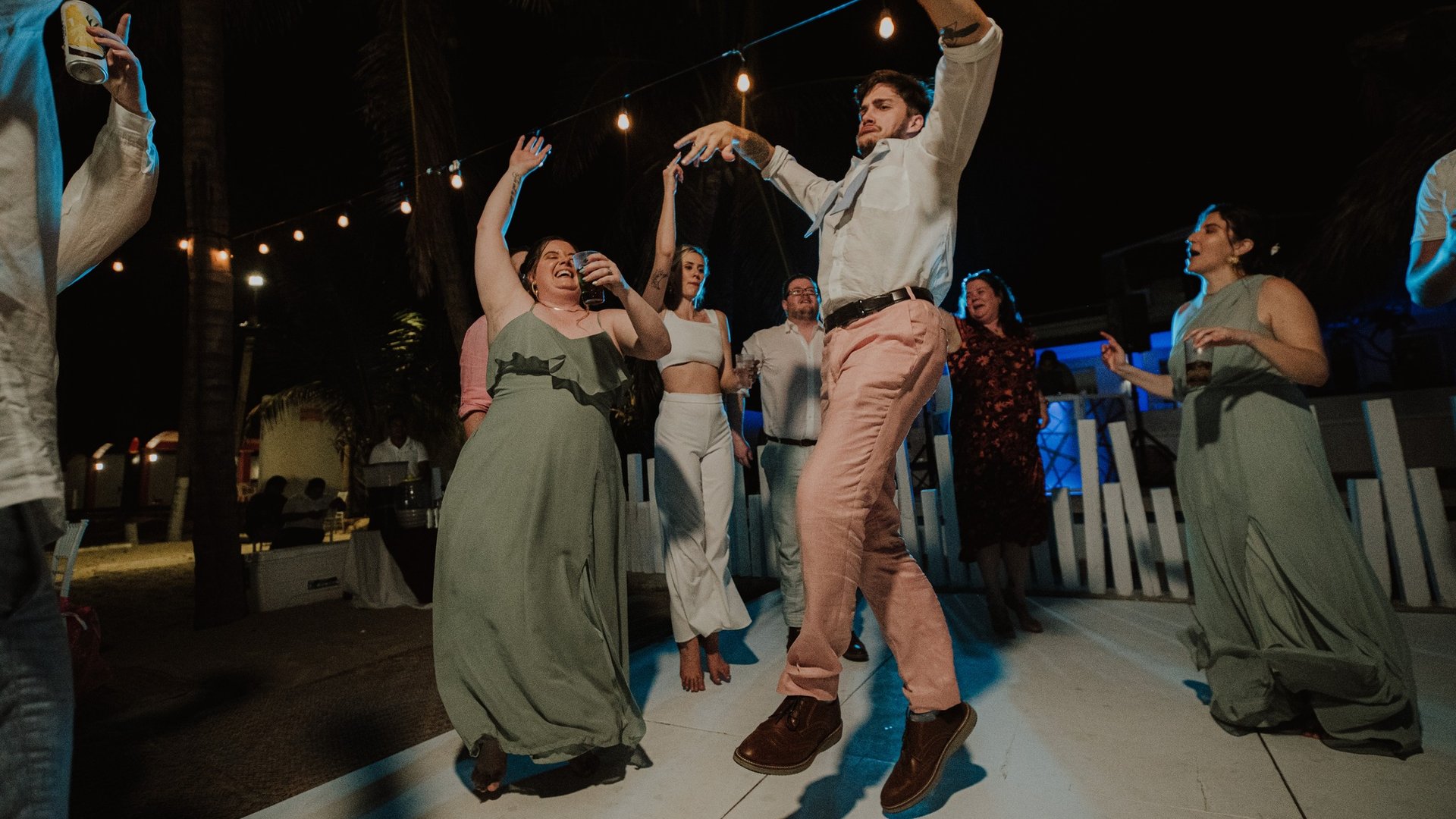 Invitados celebrando en pista de baile nocturna con luces colgantes y palmeras de fondo