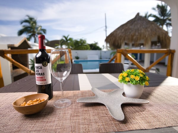 Detalle de mesa exterior con vino, copa y decoración frente a palapa y alberca al fondo