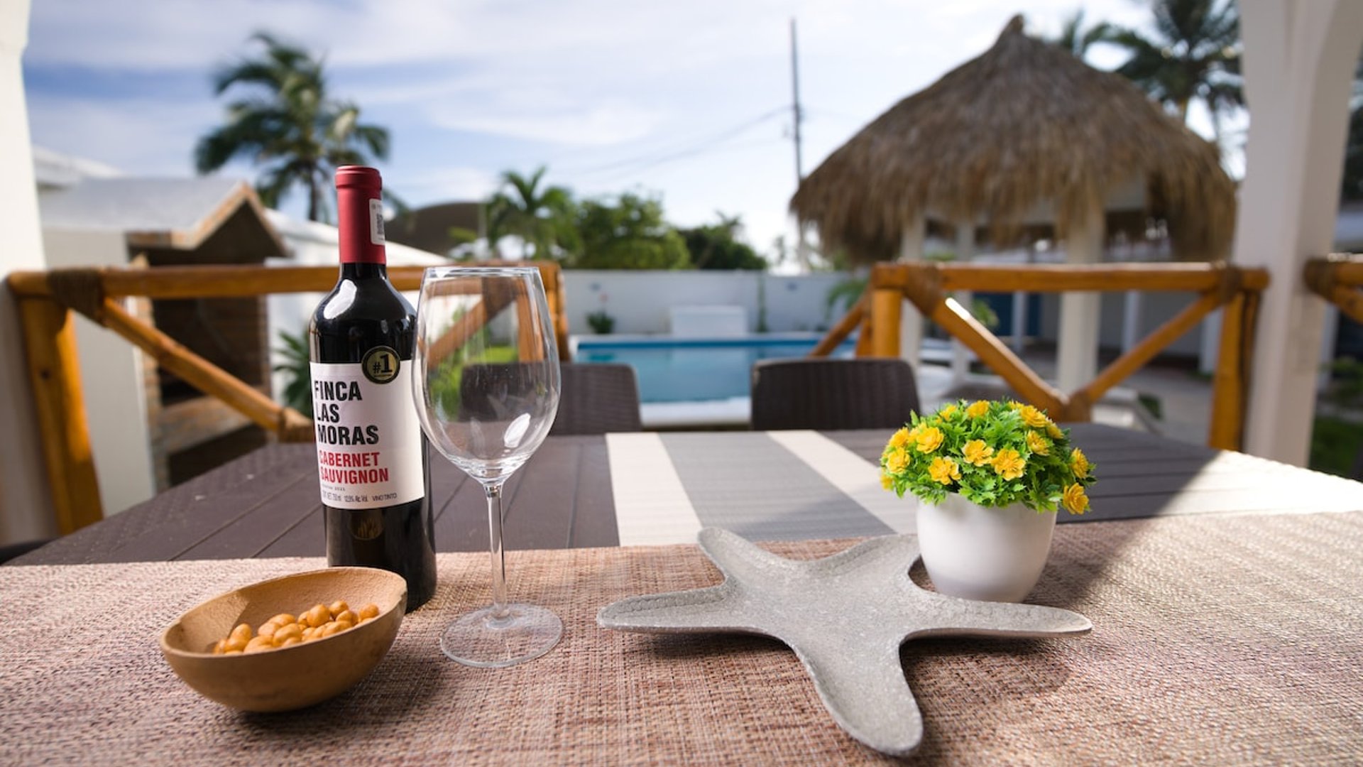 Detalle de mesa exterior con vino, copa y decoración frente a palapa y alberca al fondo