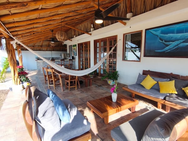 Terraza palapa con sala de madera, hamaca, comedor exterior y mural de ballenas en pared blanca