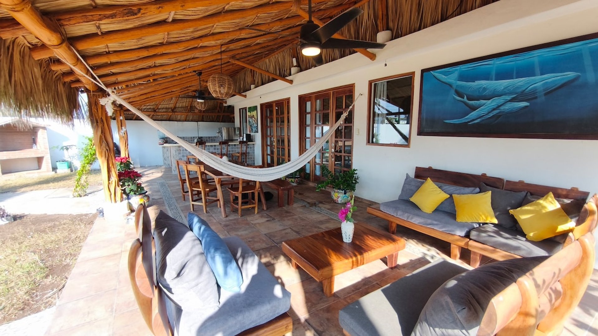 Terraza palapa con sala de madera, hamaca, comedor exterior y mural de ballenas en pared blanca