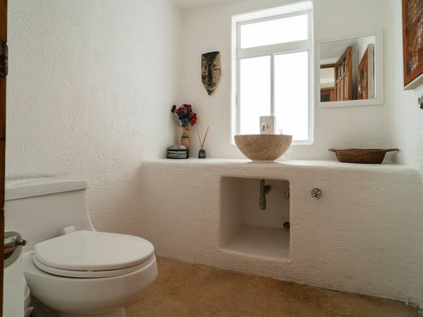 Baño blanco con lavabo de piedra sobre cubierta de mampostería, ventana cenital y detalles artesanales