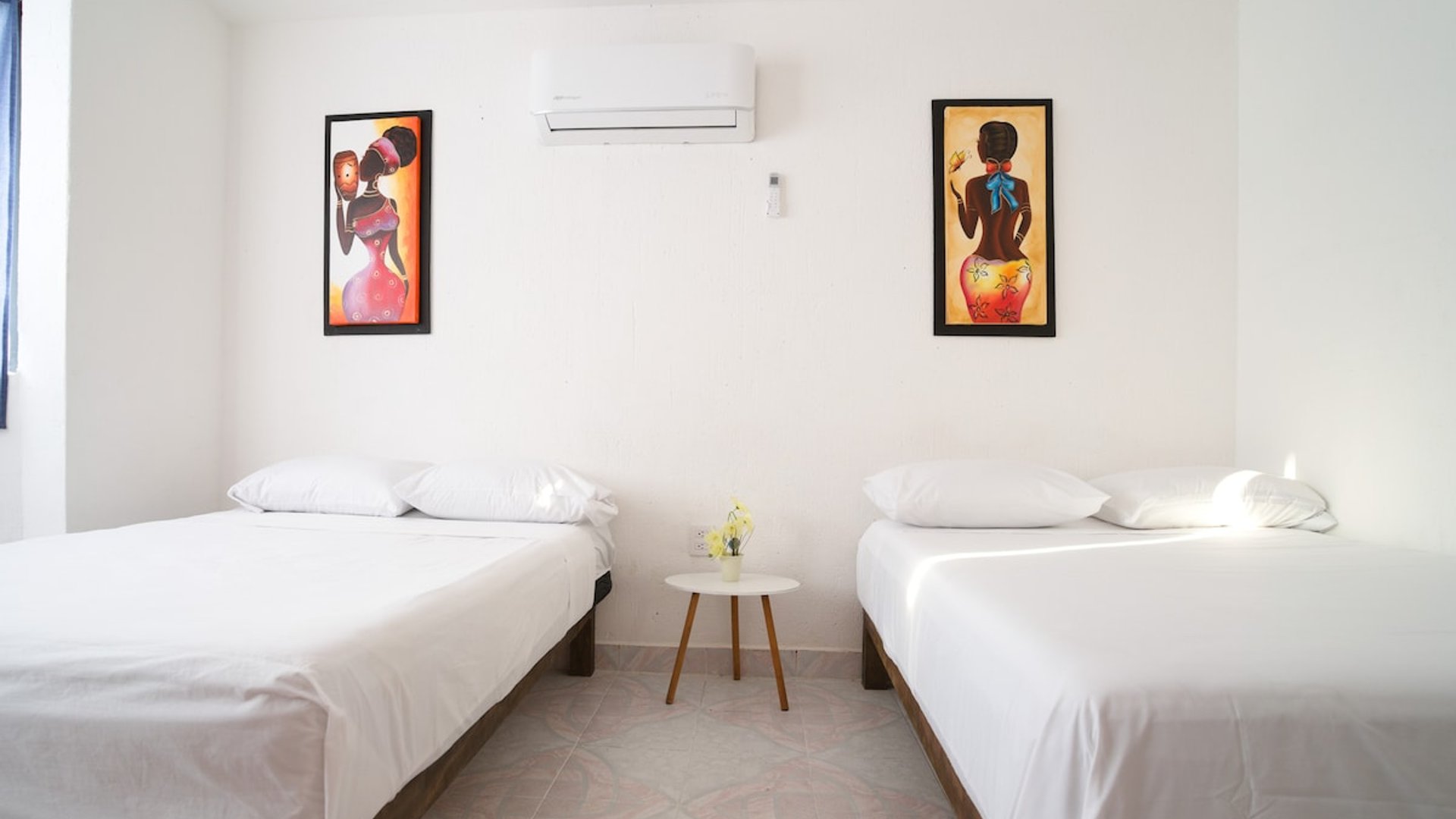 Recámara doble con dos camas individuales, sábanas blancas, aire acondicionado y cuadros coloridos en pared blanca